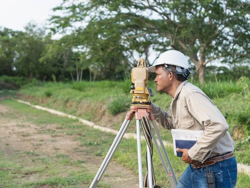 Land surveyor using a total station to measure property boundaries and site conditions in an outdoor field setting